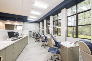 A modern dental clinic with several dental chairs facing large windows, white walls, and light wood flooring.