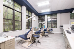 Modern dental clinic with three blue dental chairs, large windows, overhead lights, and computer workstations. The room is clean, bright, and organized with light wood flooring.