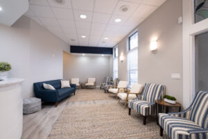 Modern waiting room with striped chairs, a blue sofa, neutral walls, large windows with blinds, and soft overhead lighting.