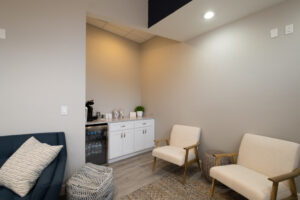 Small waiting area with two beige chairs, a blue sofa, a woven ottoman, and a kitchenette with a mini fridge, coffee maker, and snacks. Neutral walls and wood flooring.