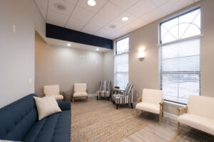 Modern waiting room with a blue sofa, striped and white chairs, a rug, and large windows with blinds letting in natural light.