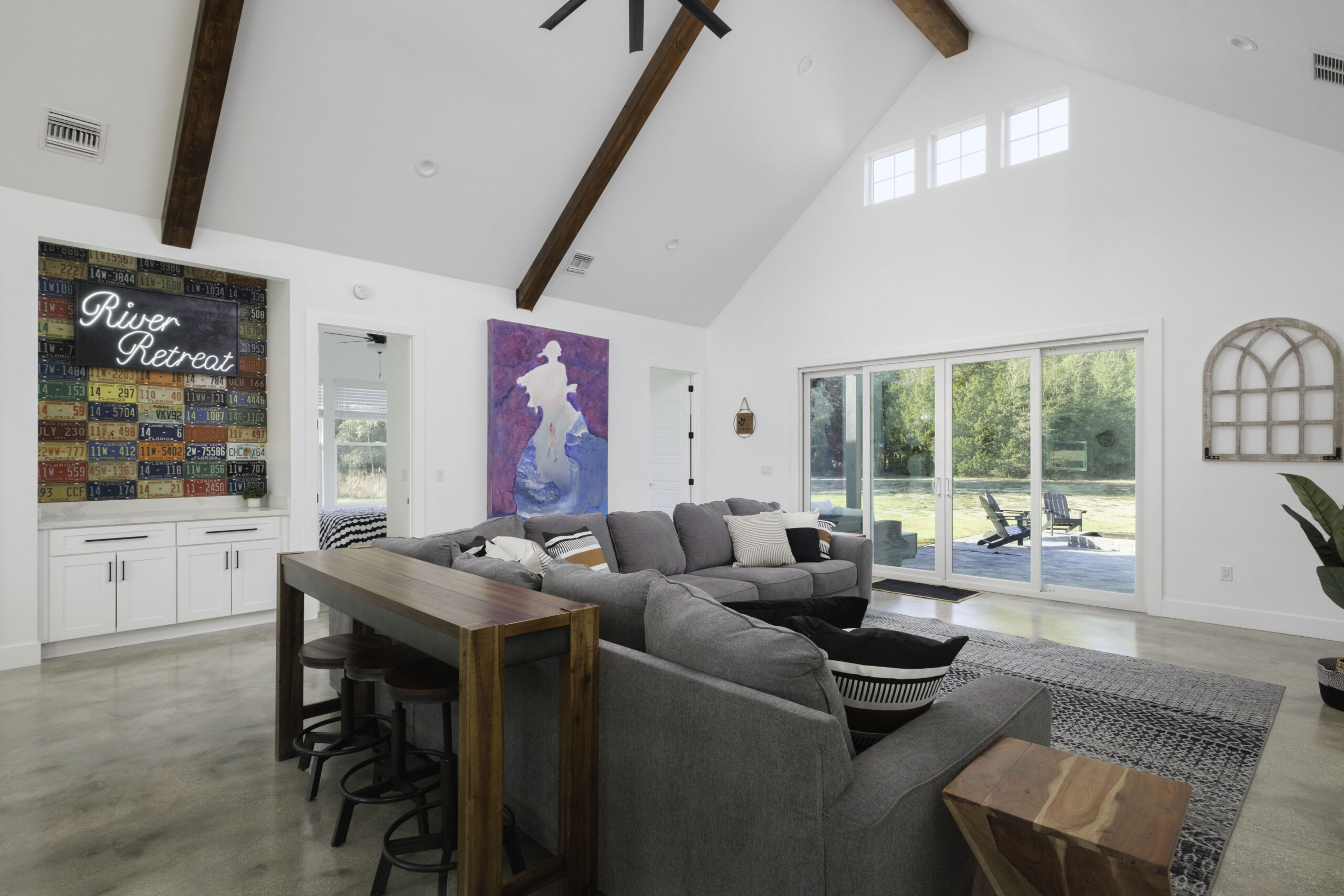A modern living room with a gray sectional sofa, wooden accents, colorful wall art, a River Retreat sign, and large sliding glass doors opening to a patio and greenery outside.