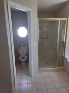 A bathroom with a glass shower, bathtub edge, and a separate toilet room with an octagonal window above the toilet.