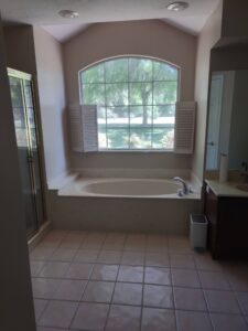 A bathroom with a large bathtub under a window, a glass shower on the left, and a vanity with a sink and mirror on the right. Sunlight filters through the trees outside.