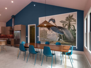 Modern dining area with a wooden table, eight blue chairs, large bird mural on a blue wall, pendant light, and open kitchen with wood cabinets and stainless steel appliances.