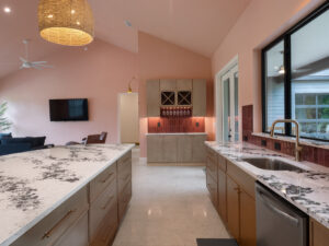 Modern kitchen with marble countertops, gold fixtures, a built-in wine rack, and a view of a living area with a wall-mounted TV and pink walls.
