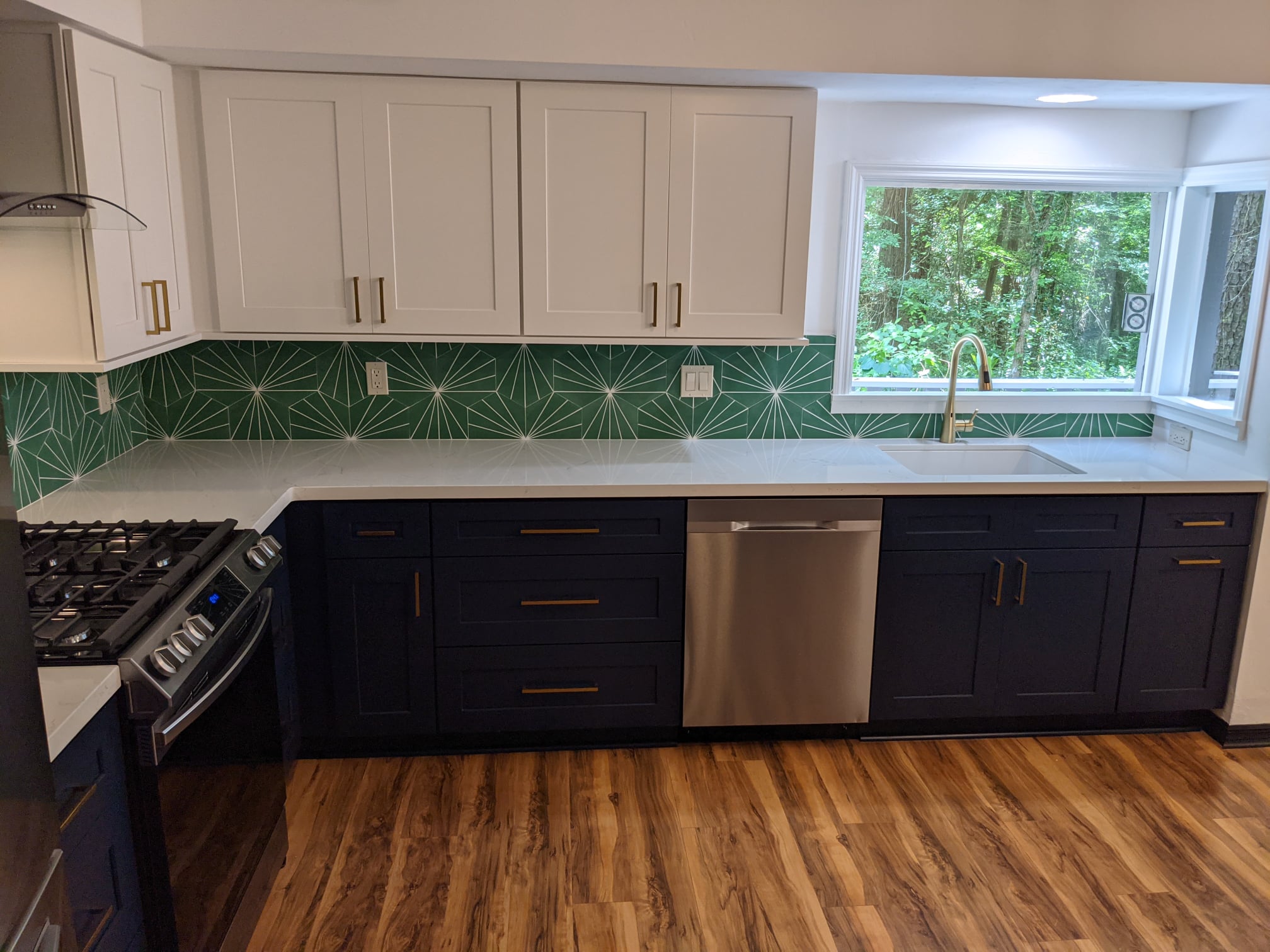 Modern kitchen with navy lower cabinets, white upper cabinets, stainless steel appliances, green geometric backsplash, and wood flooring. A large window overlooks greenery outside.