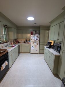 Galley kitchen with pale green cabinets, white countertops, white fridge with magnets, black appliances, and various items on counters. Overhead lighting and tile floor are visible.