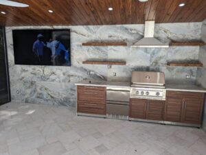 Outdoor kitchen with marble wall, wooden cabinets, stainless steel grill, range hood, floating shelves, and a wall-mounted TV displaying people.