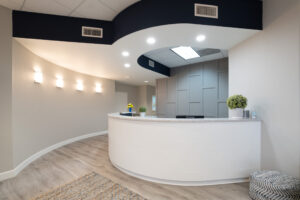 Modern, curved reception desk in a brightly lit office lobby with light wood flooring, gray walls, wall lights, and small potted plants on the counter.