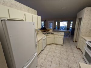 A kitchen with white cabinets, white appliances, beige countertops, tile floor, and wallpapered walls. A dining area with large windows is visible in the background.