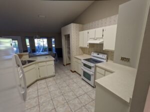 A kitchen with white cabinets, tile floor, electric stove, and fridge. A peninsula counter separates the kitchen from the open dining and living area with large windows.