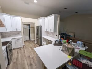 Modern kitchen with white cabinets, stainless steel appliances, and wood-style flooring; countertop has various household items and supplies.