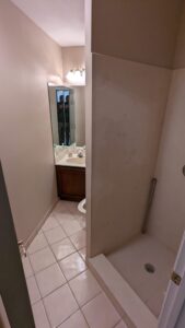 Small bathroom with a tiled floor, vanity sink with mirror, overhead light, and a walk-in shower with a grab bar.