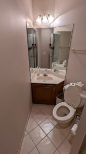 Small bathroom with a single sink vanity, large wall mirror, toilet, towel rack, waste bin, and tile floor under overhead lighting.