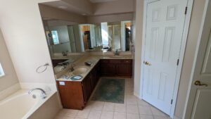 Bathroom with a double sink vanity, large mirror, white tile floor, bathtub, and a closed door. A green mat is placed in front of the sinks.