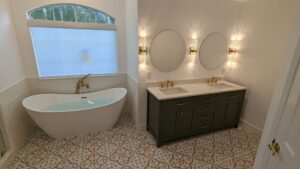 Modern bathroom with a freestanding tub, green double-sink vanity with gold fixtures, round mirrors, and decorative tile floor.