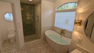 Modern bathroom with patterned floor, glass shower, freestanding tub with water, toilet, and large frosted window.