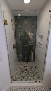 Glass shower with geometric dark green tile accent wall, brass fixtures, white subway tiles, built-in shelf, and pebble stone floor.