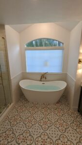 A modern bathroom features a white freestanding bathtub, gold fixtures, patterned tile floor, and a large window with a shade.