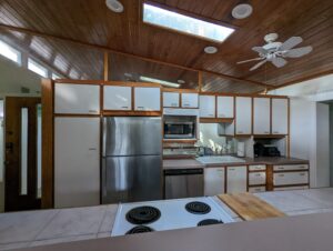 Modern kitchen with wood-paneled ceiling, white cabinets, stainless steel refrigerator, microwave, sink, stove, and skylight. Countertops feature small appliances and a cutting board.