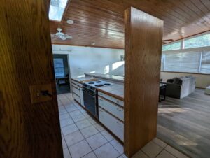 View of a kitchen with a stove and white cabinets, adjacent to a living room area with a couch, large windows, and wood-paneled ceiling.