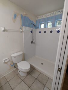 Bathroom with a toilet and walk-in shower; partially removed wallpaper on walls, white tiles with blue accents, and tiled floor.