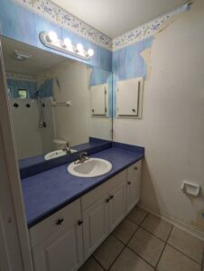Bathroom with blue countertop, white sink, large mirror, and partially stripped wallpaper; light fixture above mirror and tiled floor visible.
