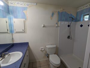 Bathroom with blue countertop, sink, toilet, shower, wall cabinet, and partially peeled wallpaper on the upper walls.