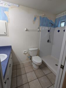 A small bathroom with peeling wallpaper, a toilet, a single sink vanity, a shower with white tiles, and a tiled floor.