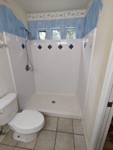 Shower with white tile and blue diamond accents, adjacent to a toilet. Three small windows above let in natural light. Blue and floral wallpaper borders the top of the walls.