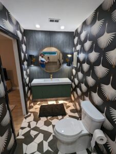 A bathroom with crane-patterned wallpaper, geometric tile floor, a toilet, a floating vanity with a round mirror, and a person taking a photo reflected in the mirror.
