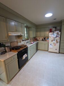 A kitchen with pale green cabinets, white countertops, a black dishwasher, a white refrigerator with magnets, and various items on the counters.
