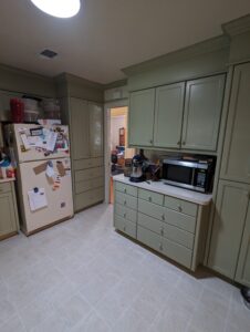 A kitchen with green cabinets, a refrigerator covered in magnets and papers, a microwave, mixer, and various items on the countertops.