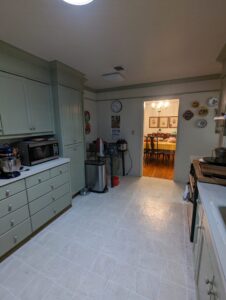 A kitchen with light green cabinets, white countertops, appliances, and a view into a dining room with a lit chandelier and framed art on the wall.