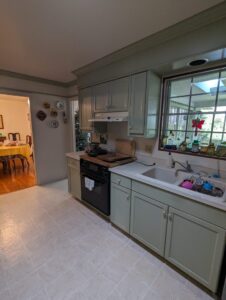 A kitchen with green cabinets, a black stove, white countertop, sink with dishes, and a window above the sink. Dining room visible through the doorway.
