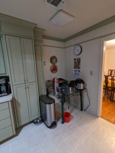 Corner of a kitchen with green cabinets, a microwave, two trash bins, small appliances on a table, a wall clock, and posters; a dining room is visible through an open doorway.