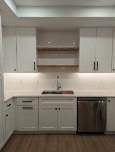 Modern kitchen with white cabinets, wood shelves, stainless steel dishwasher, under-cabinet lighting, white tile backsplash, and wood flooring. Sink is centered below the shelves.
