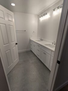 A bathroom with gray floor tiles, white double sink vanity, wall-mounted light fixtures, and an open white door.