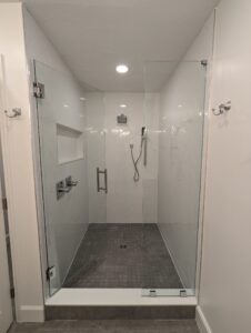 Modern shower with glass doors, white tile walls in a herringbone pattern, overhead rain shower, handheld shower, built-in shelf, and dark floor tiles.