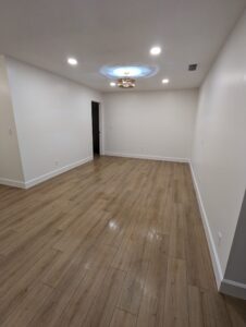 Empty room with light-colored wooden flooring, white walls, recessed ceiling lights, and a small chandelier at the center. The room is unfurnished and appears newly renovated.