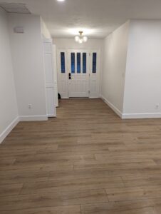 A hallway with light wood flooring, white walls, a ceiling light fixture, and a white front door with glass panels.