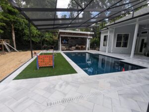 Modern backyard with a marble-tiled patio, rectangular pool, large Connect Four game on artificial grass, and covered outdoor seating area with TV.
