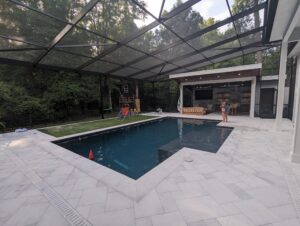 Enclosed backyard pool area with lawn, patio, playset, lounge furniture, and a person standing near the pool under a covered section. Trees and fencing surround the outdoor space.