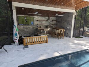 Outdoor lounge area by a pool with a grill, dining table, sofa, and TV displaying golf, under a wooden ceiling with fans. Towels hang near the sofa; greenery is visible in the background.