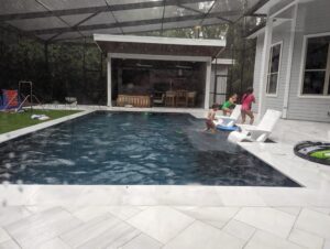 Three children play by a backyard pool during rainfall, with outdoor furniture and toys visible around the patio area.