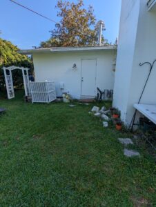 Small white building with a closed door, utility meters, and scattered stones; grass lawn in front, white trellis and various plants along the sides.