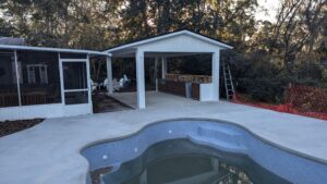 An empty in-ground pool is next to a covered outdoor kitchen area and patio, with trees and construction materials in the background.