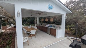 Outdoor covered kitchen and dining area with a long table, white chairs, built-in grill, counter space, ceiling fans, and a decorative blue crab on the gable.