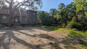 A section of dirt yard near a gray house is marked off with yellow caution tape and orange barriers; trees and greenery surround the area.